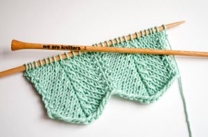 swatch of green yarn on wooden straight needles size US 5; the top needle is engraved with the words "we are knitters"