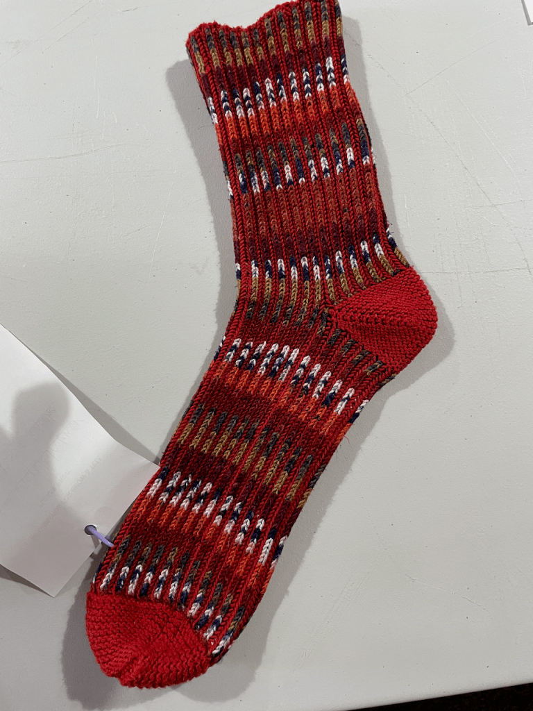 photo of one Biscotte Brioche sock in red (main color, solid heel and toe, with variegated black/brown/white/grey in between the brioche ribs)