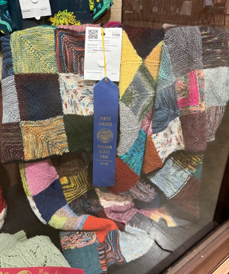 photo of Marcia's mitre-square multi-colored blanket hanging in a display case with a blue ribbon pinned and hanging in front of it